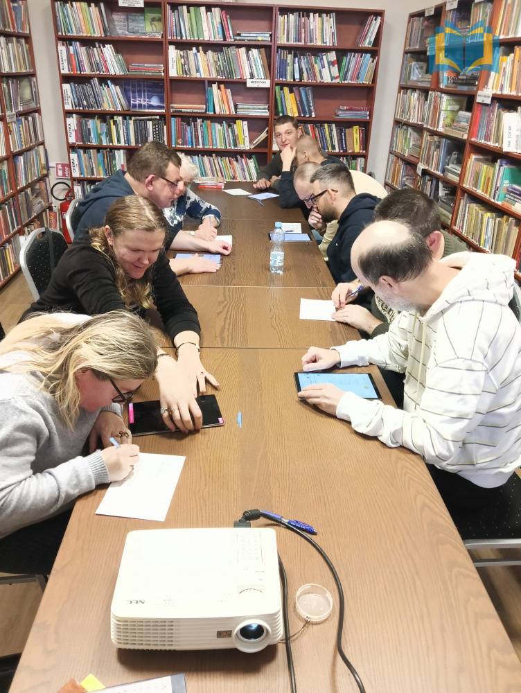 Zdjęcie: Dorośli uczestnicy WTZ za pomocą tabletów wyszukują informacje o zbiorach bibliotecznych. Siedzą przy długim brązowym stole w bibliotece.